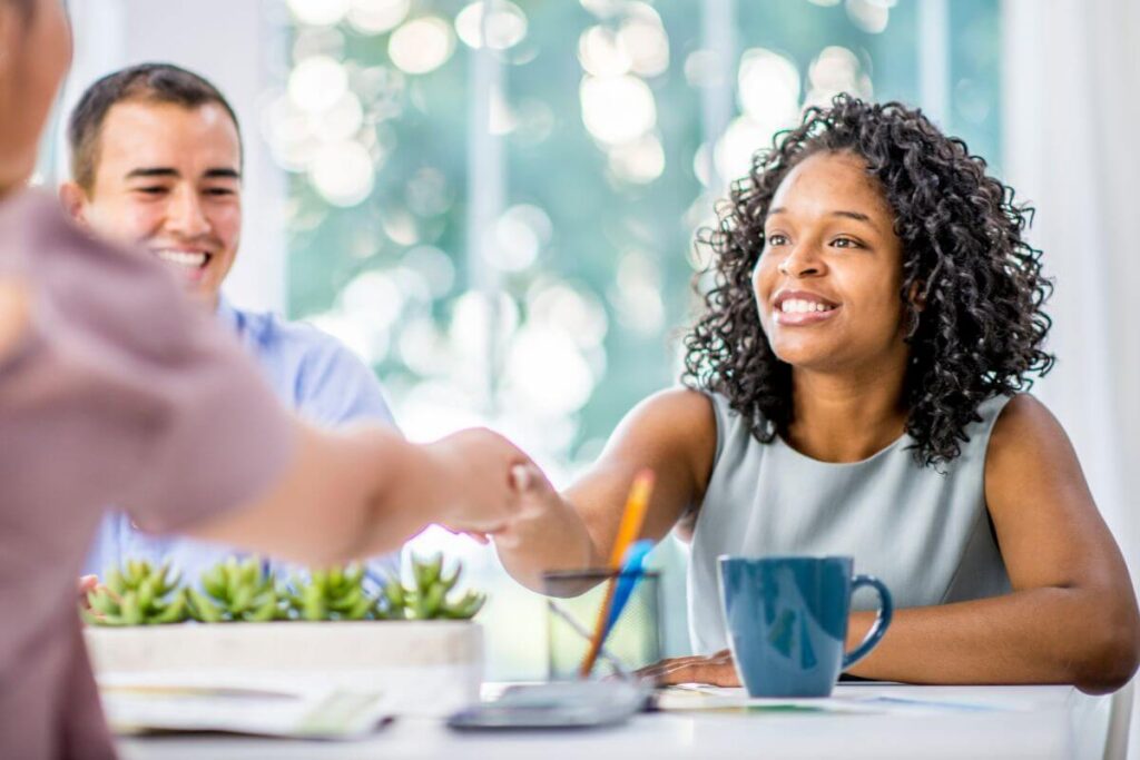 three people sit at a table together and politely shake hands to show proper etiquette when greeting someone