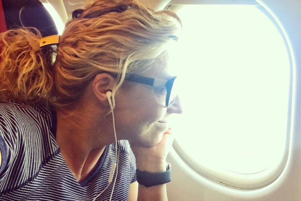 woman looking out an airplane window with earplugs in listening to the best adventure songs