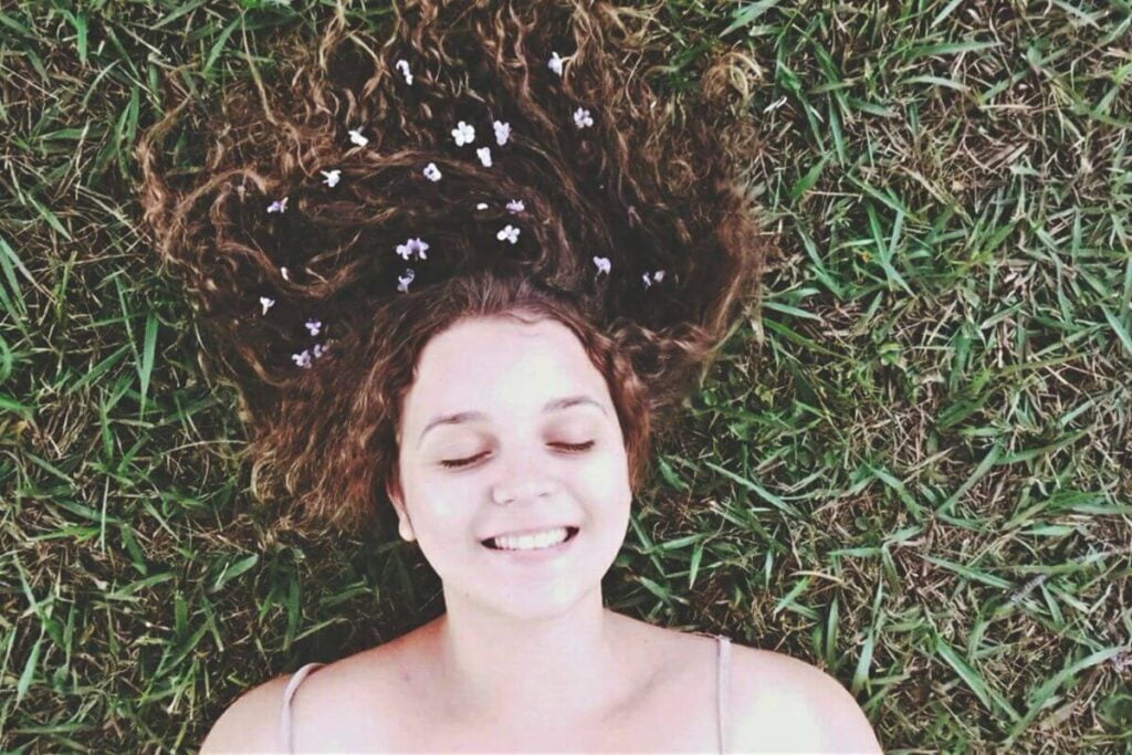 a young dark haired happy woman lays in the grass with her hair on top of her head decorated with flowers