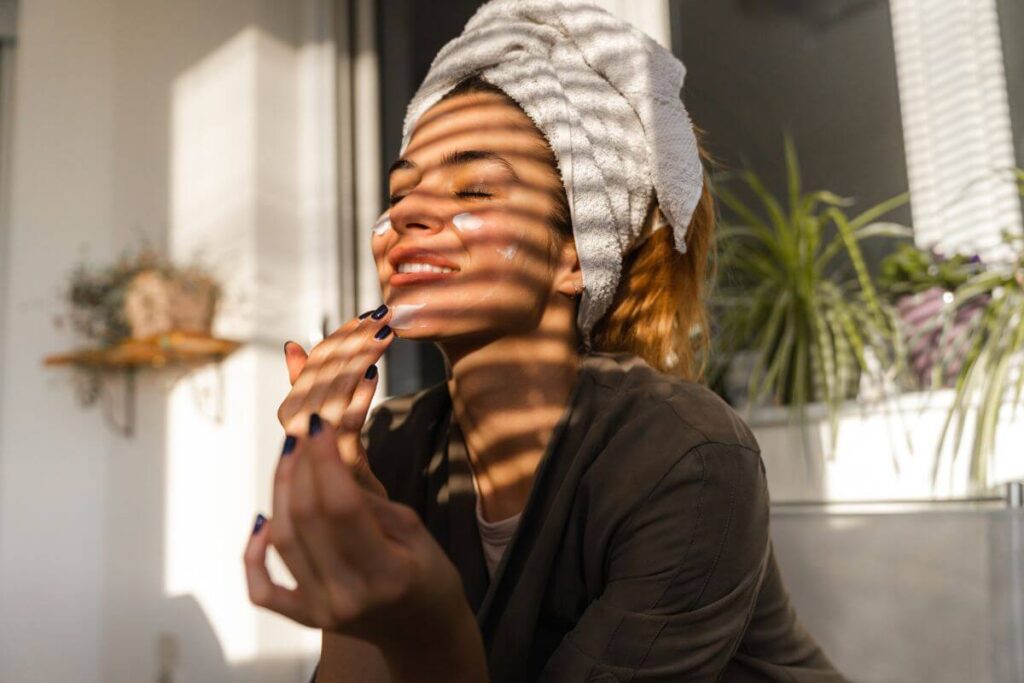 a young woman sits in the sun in a robe and gives herself a facial for self care
