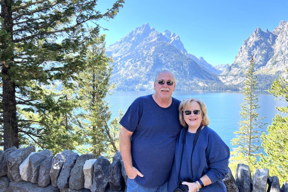 a middle aged couple stands againsts a crystal blue lake at the base of mountains after using travel affirmations to plan a trip of their lifetime