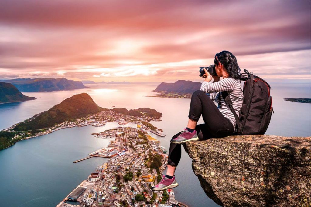 a young adventurous women taking photos sits on a large boulder over looking a city on the water