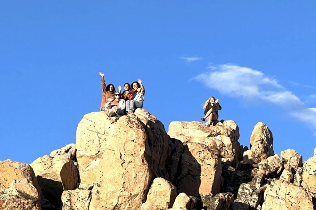 five young women sit on boulders at the summit of a mountain after using travel affirmations to give them confidence