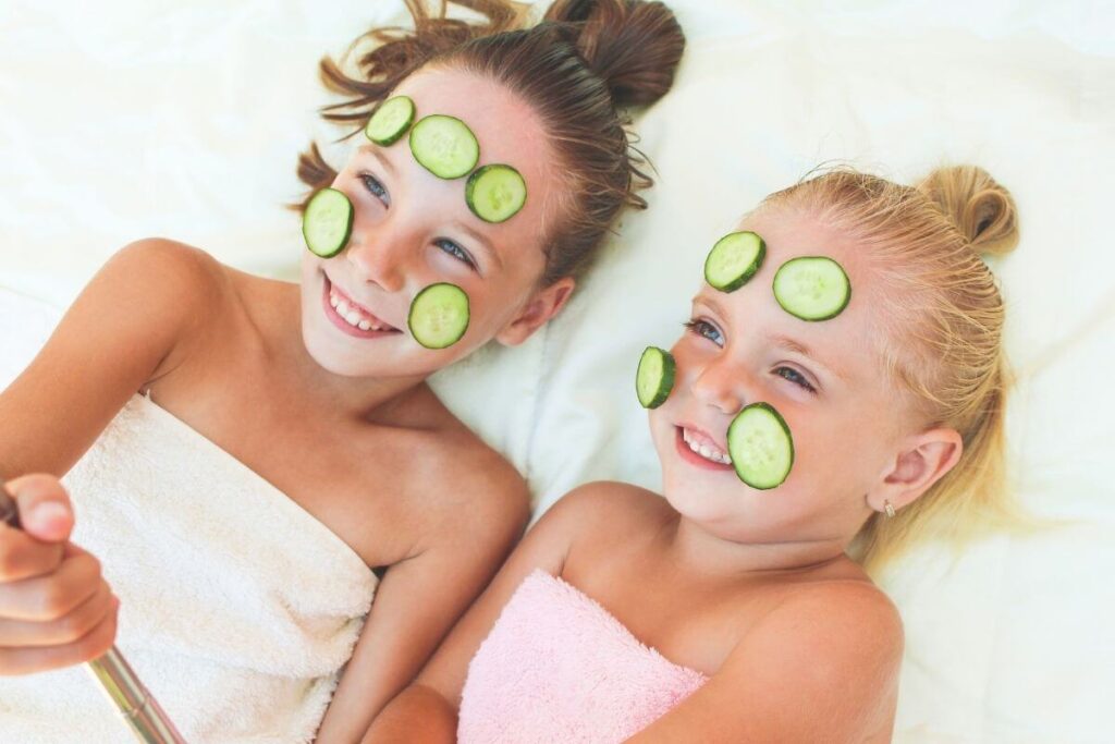 two young girls lay on a bed wrapped in towels with cucumber slices on their faces for a fun spa day