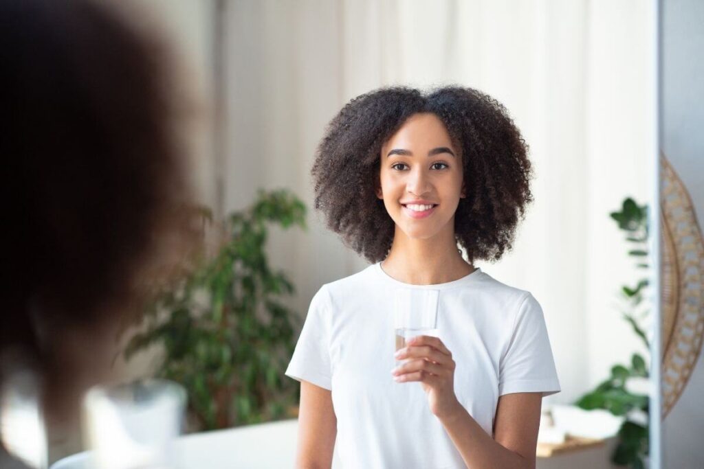 beautiful young black woman holds a tall glass of water and smiles pleasantly about her emotional self care routine
