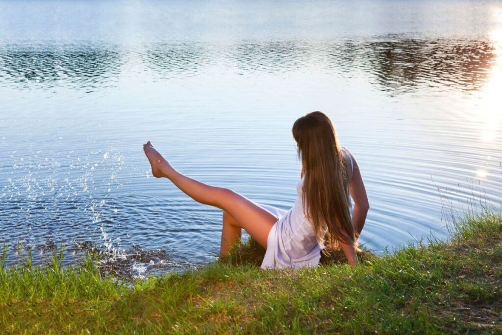 a woman with long brown hair falling to her waist sits along the edge of a body of smooth glistening water with one foot playfully flicking water in the air