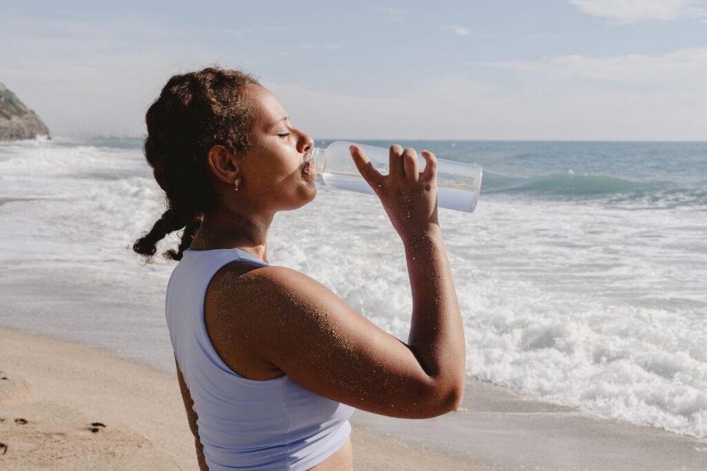 a young woman stands on a beach and drinks her water after speaking her affirmations for health and wellness