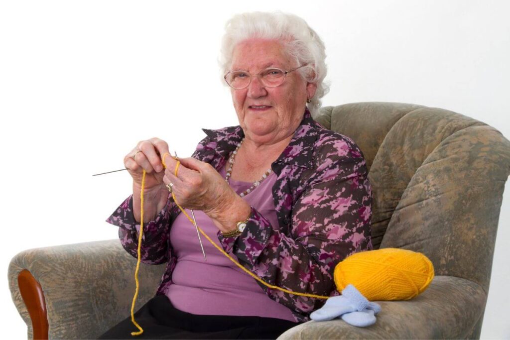 an elderly women sits in a plush chair in her home with her knitting