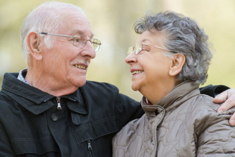 an elderly couple happily smile at each other outside on a beautiful day