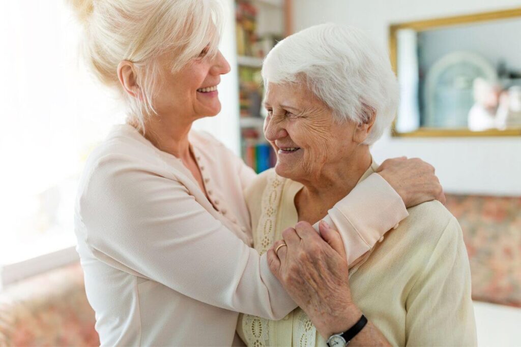 elderly woman smiling in her new home with her adult daughter hugging her