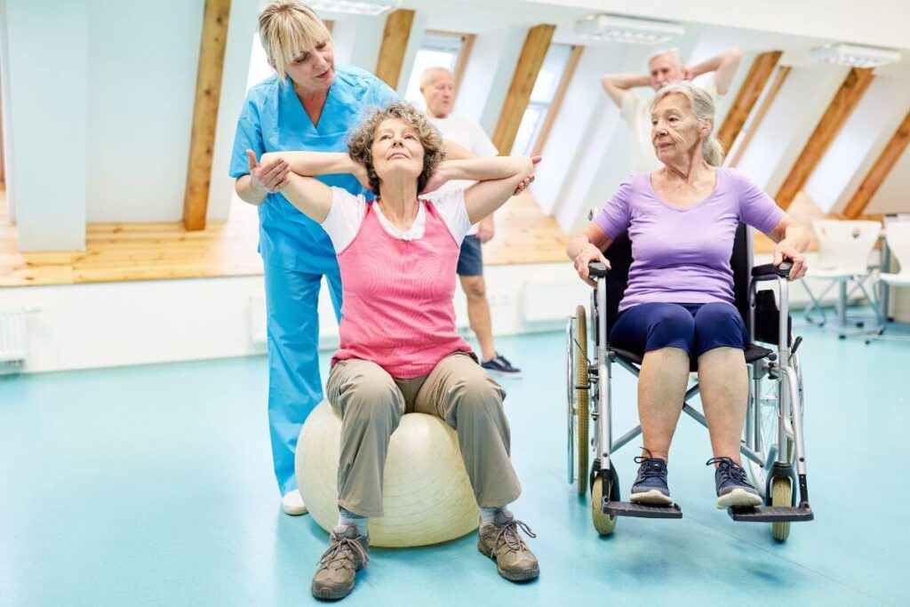 a physical therapist assists clients with mobility issues in a small class of four with one elderly woman in a wheelchair