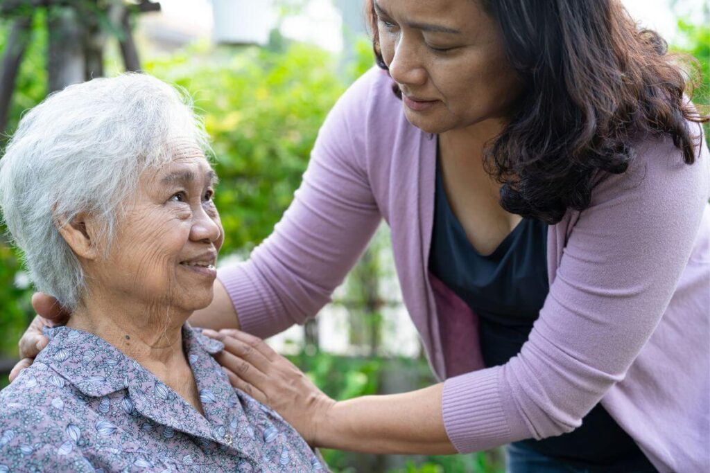 Asian woman bends over her mother appearing to be feeling trapped caring for elderly parents