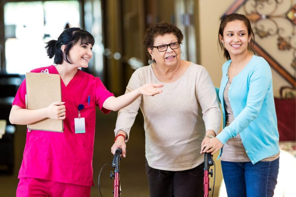 an elderly woman and her daugher are shown around her new assisted living facility by a cheerful member of the nursing staff