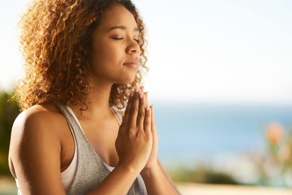 a beautiful brown skinned woman meditates outside along the water's edge