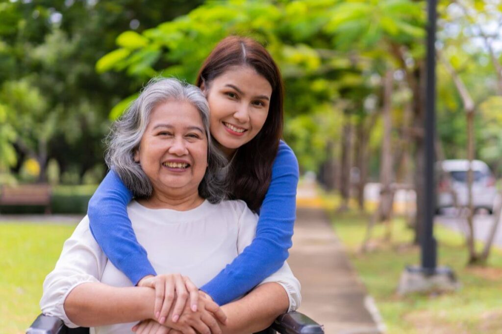 a loving woman wraps her arms around her elderly mother who is sitting in a wheelchair