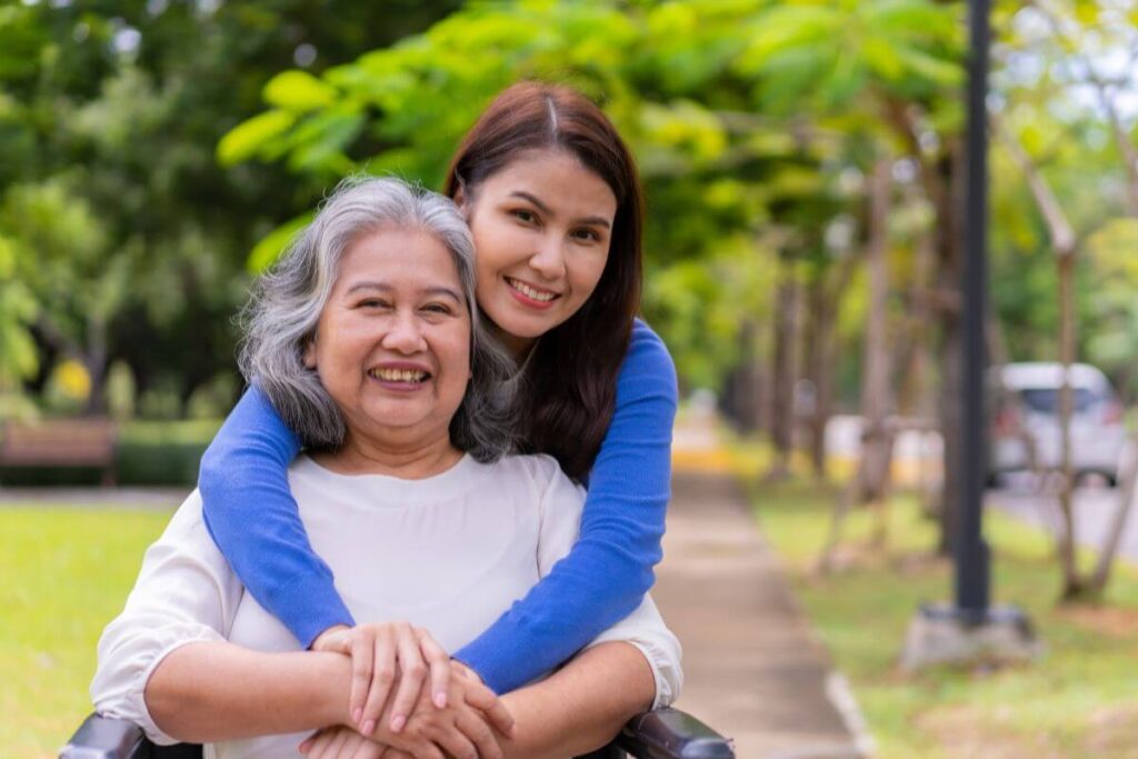 a loving woman wraps her arms around her elderly mother who is sitting in a wheelchair