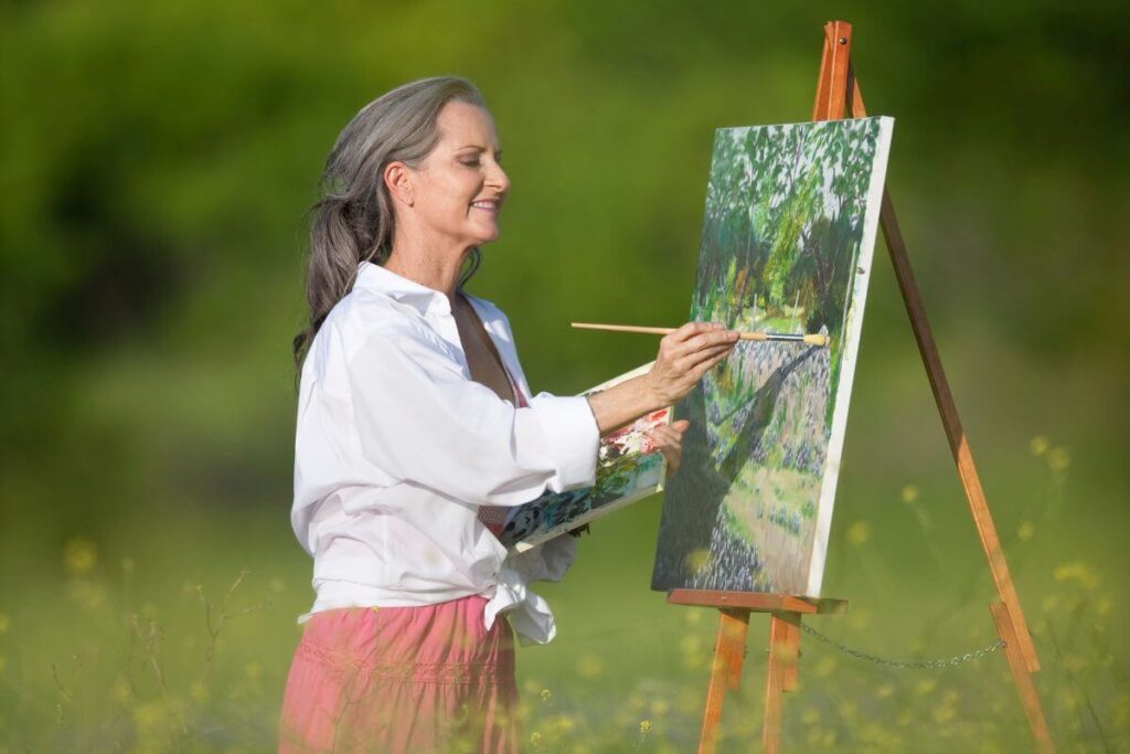 a happy middle aged women stands in a field of tall grass as she paints a canvas picture