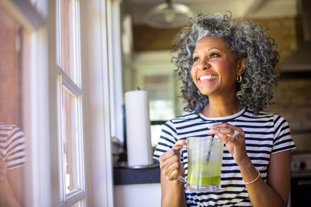 a beautiful black women with silver hair looks out a window in her home reflecting about finding a purpose in life after 50
