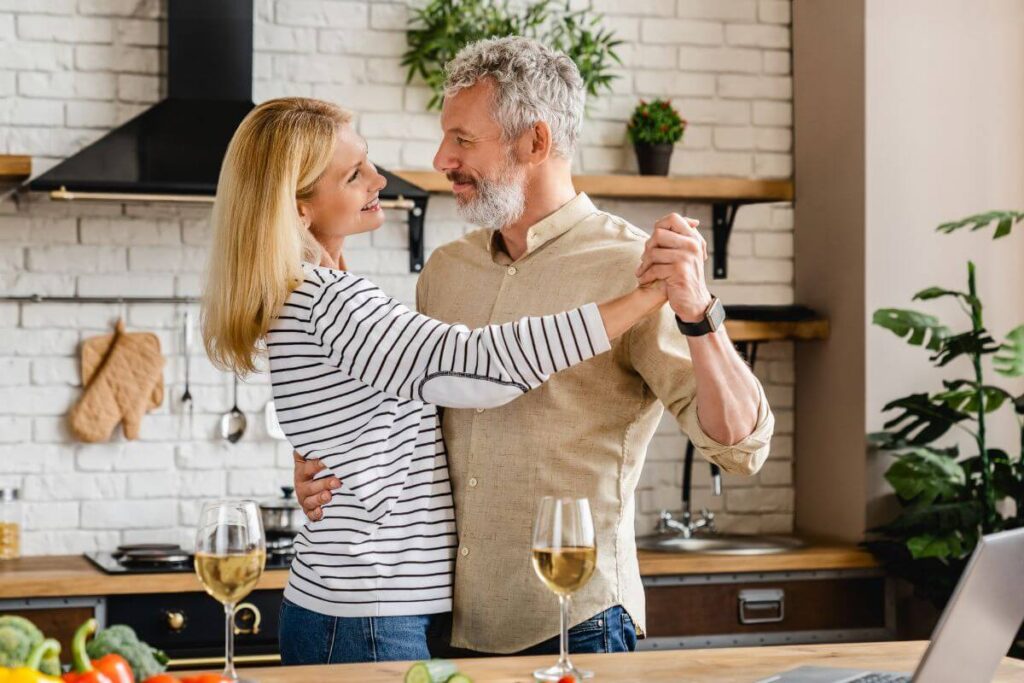 a middle aged couple shows how relationships improve as you age while they dance in their kitchen with wine glasses sitting on the counter