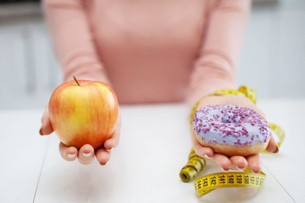 a close up of female holding an apple in one hand a donut wrapped in a tape measure in the other hand as if she is weighing her options