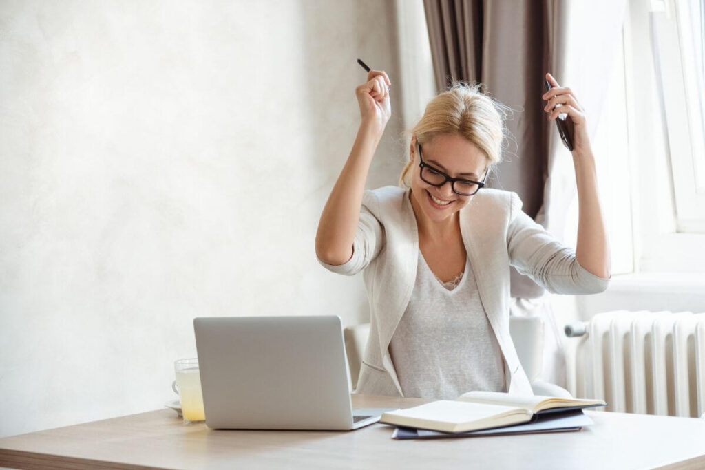 a middle aged woman celebrates her work accomplishment at her desk behind her laptop and books