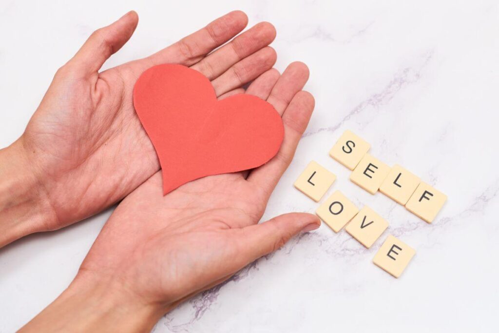 top view of hands holding a red heart cutout heart and small tiles that say self love