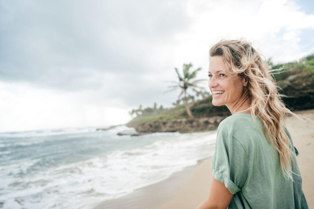 a smiling middle-aged woman sits on a beach alone learning to nourish herself with self love