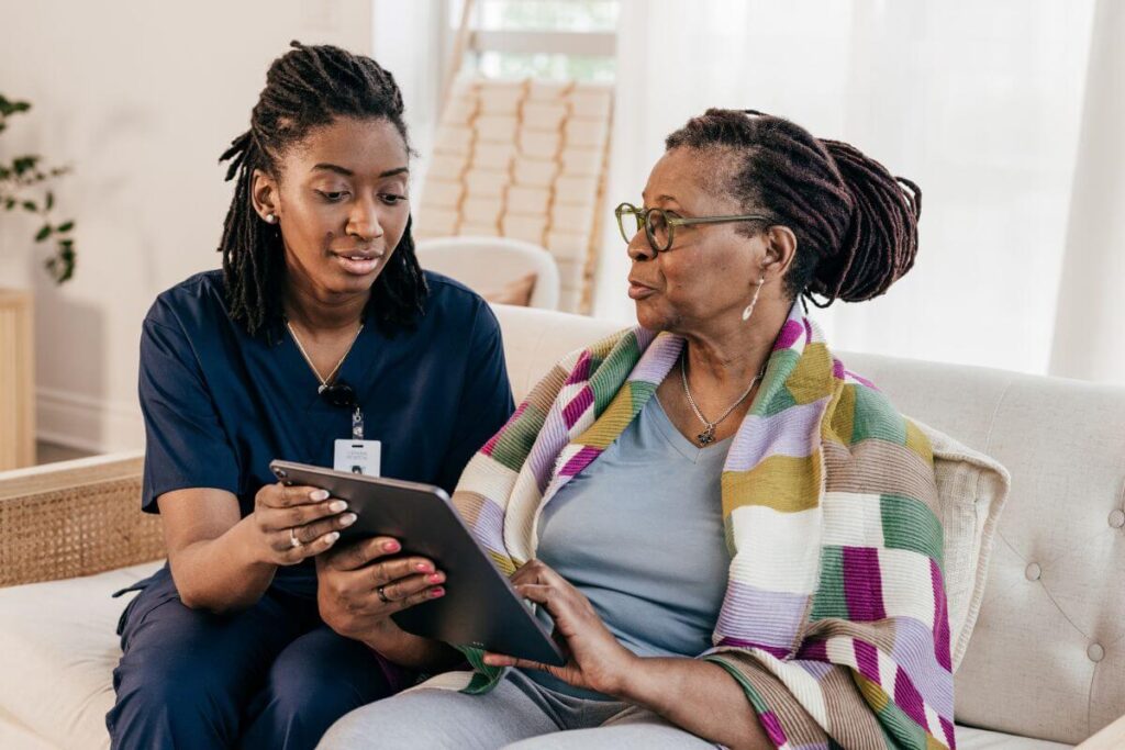 a hospice nurse sits on a sofa with a patient who has a colorful wrap on her shoulders