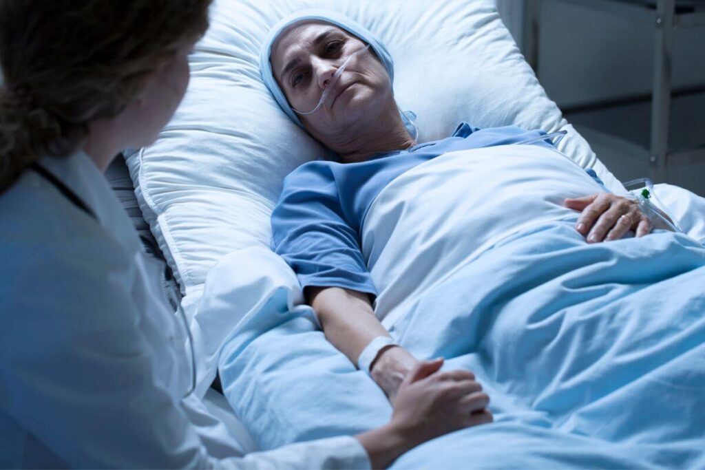 a very ill hospice patient lies in bed and someone sits next to her holding her hand