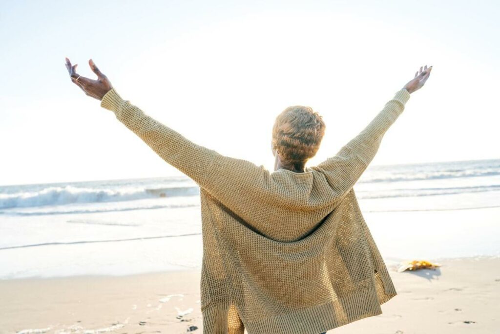 middle aged woman freely stands on the shoreline of a beach with her arms in the air