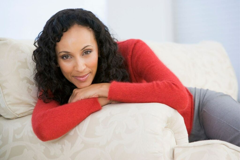 a beautiful dark skinned middle aged woman wearing a red sweater sits on a sofa and smiles at the camera
