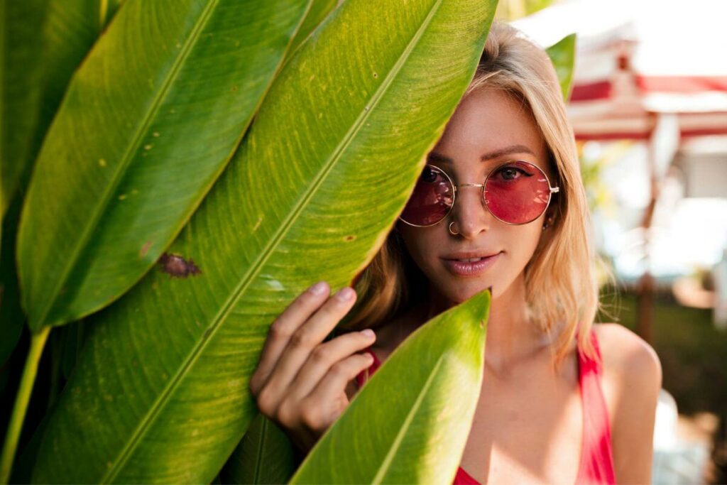 young woman wearing round sunglasses peeking from behind a large leaf as she reflects on Brene Brown Gifts of Imperfection Quotes