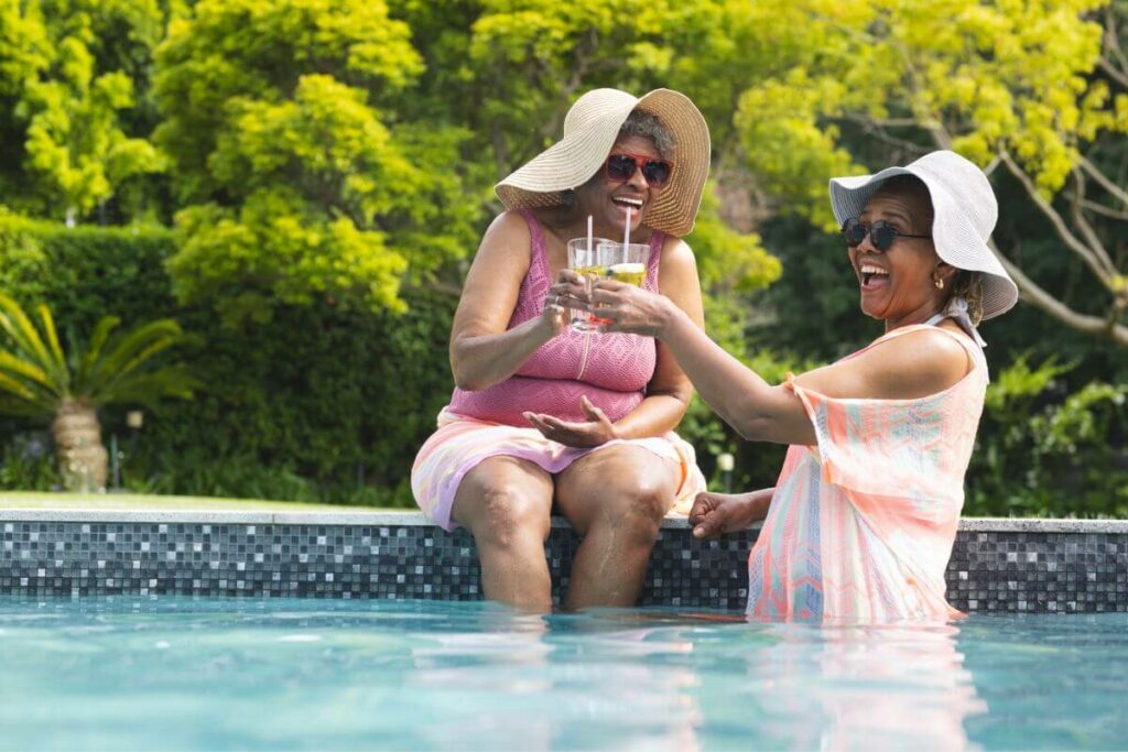 two senior women having a blast together, one sitting on the edge of a pool and one in the pool, both with colorful bathing suits on and drinks in their hands