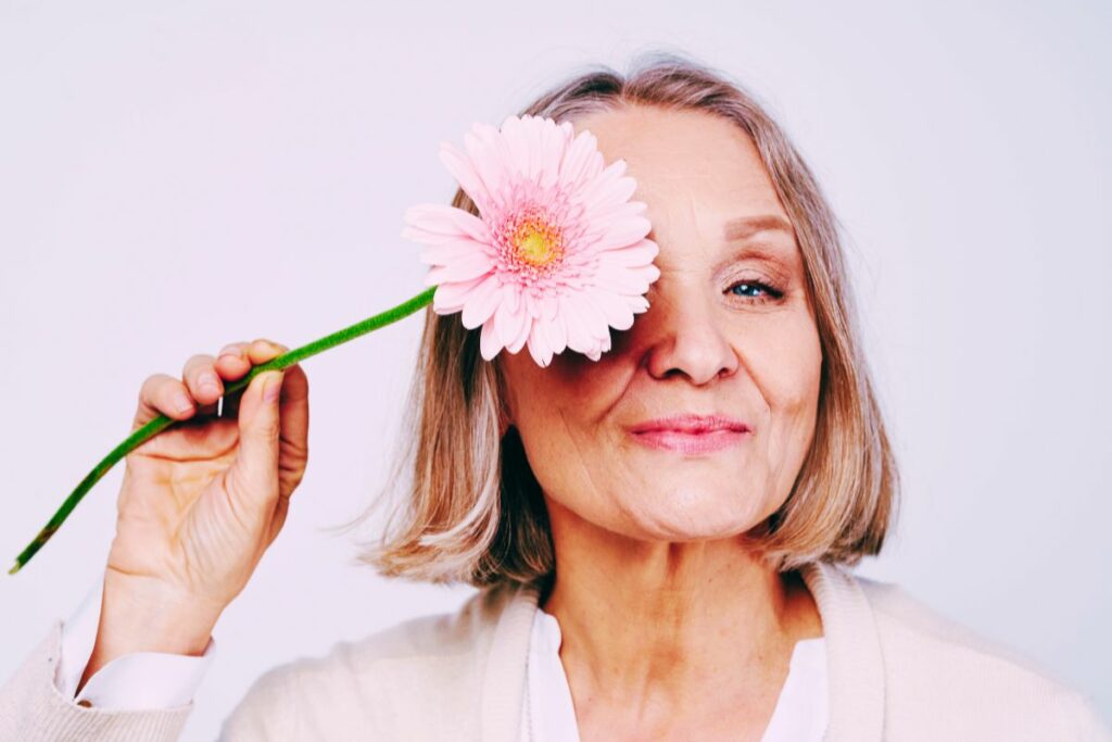 close up of a happy middle aged woman holding a pink daisy in front of her right eye because she's been inspired by let yourself bloom quotes