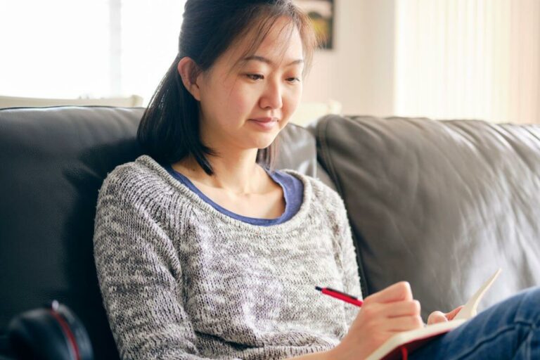 middle aged Asian woman sits on a sofa and uses personal growth journal prompts to write in her journal.