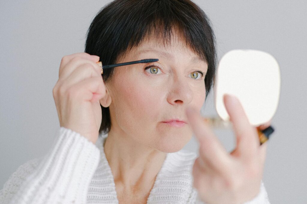 A senior woman in a cozy sweater applying mascara with a focused expression indoors.Anna Shvets