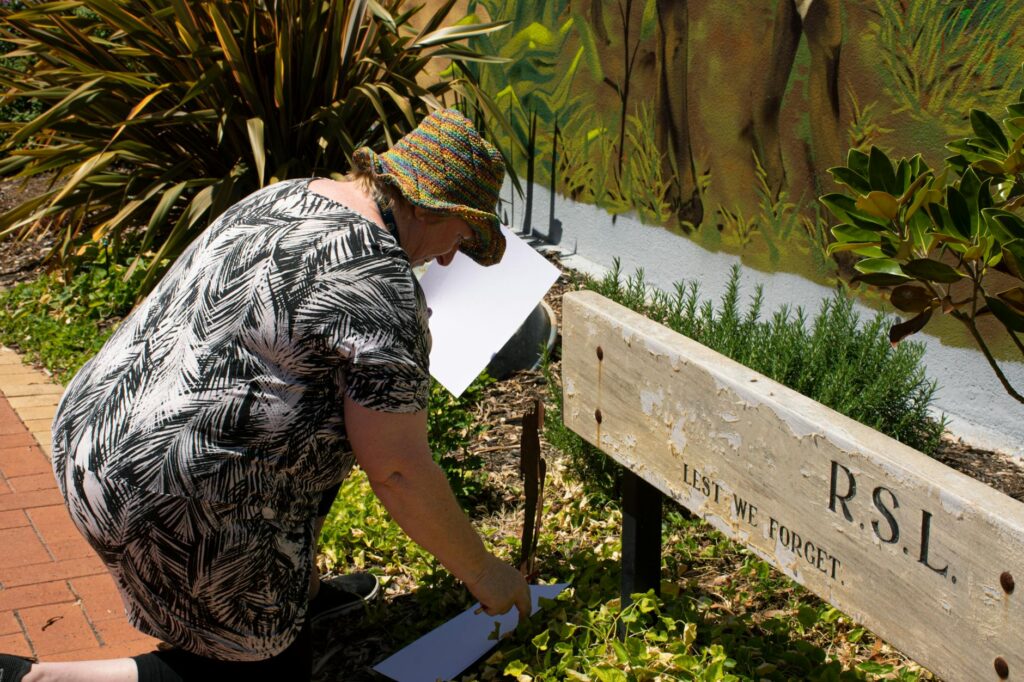 A senior woman painting outdoors near an RSL memorial bench.