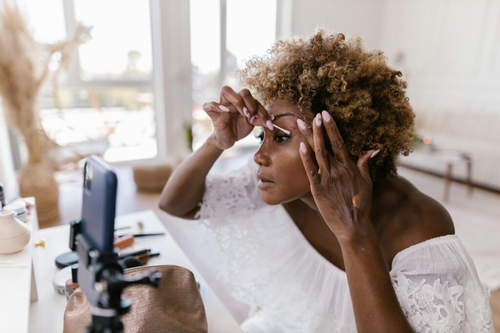 A woman applying makeup while recording her beauty routine on smartphone indoors.