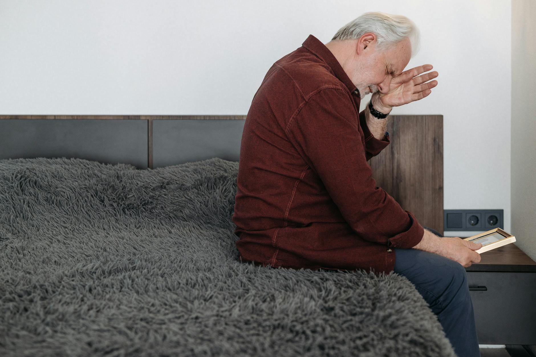 An elderly man sits on a bed in tears holding a picture frame depicting bereavement.Pavel Danilyuk