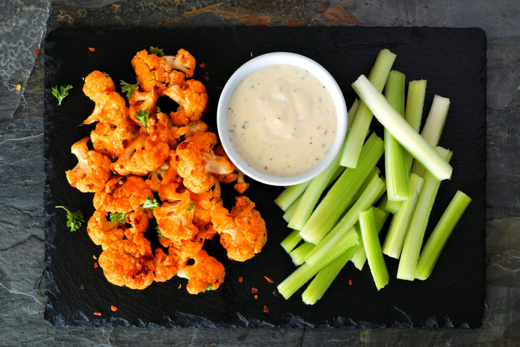 Cauliflower buffalo wings with celery and ranch dip. Top view on a slate serving platter. Healthy eating, plant based meat substitute concept.