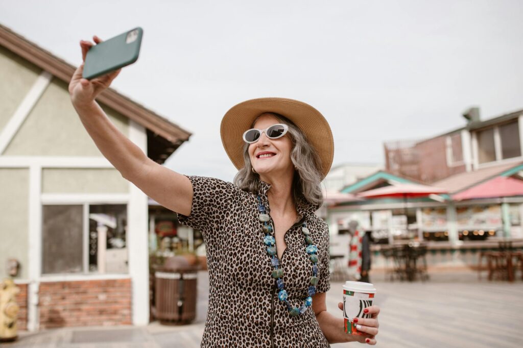 Cheerful senior woman wearing sunglasses and a hat, taking a selfie with a smartphone outdoors.