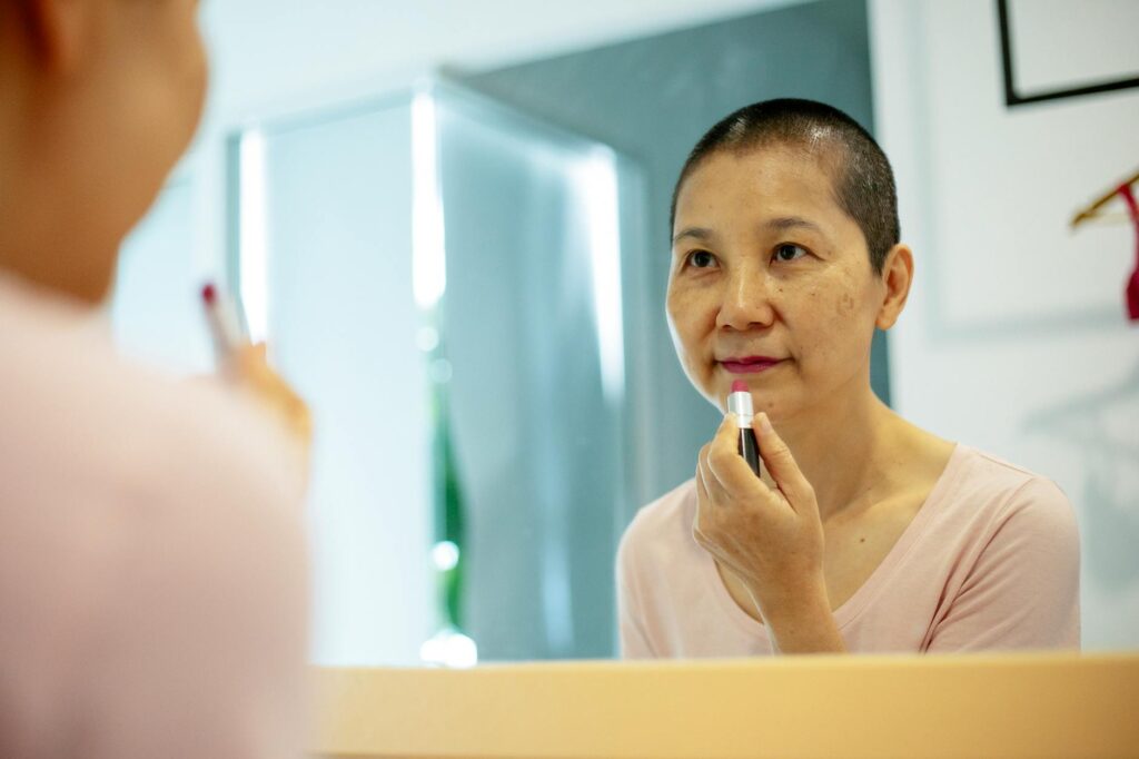 Confident middle age ethnic female in casual outfit applying purple lipstick on lips while looking at mirror reflection in bright apartment