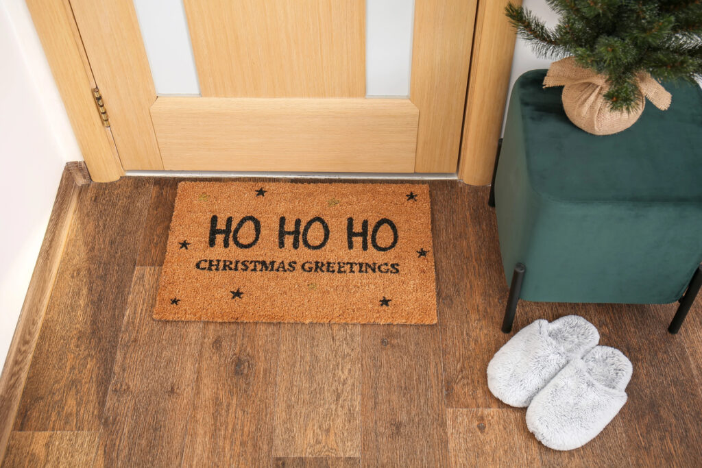 Mat with Christmas greeting, slippers and pouf near wooden door