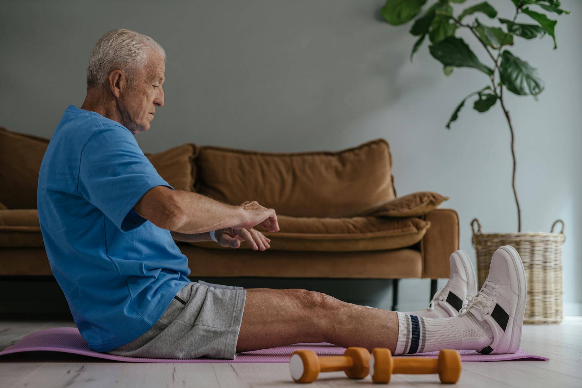Elderly man in blue shirt exercising on a yoga mat indoors with dumbbells in view.
