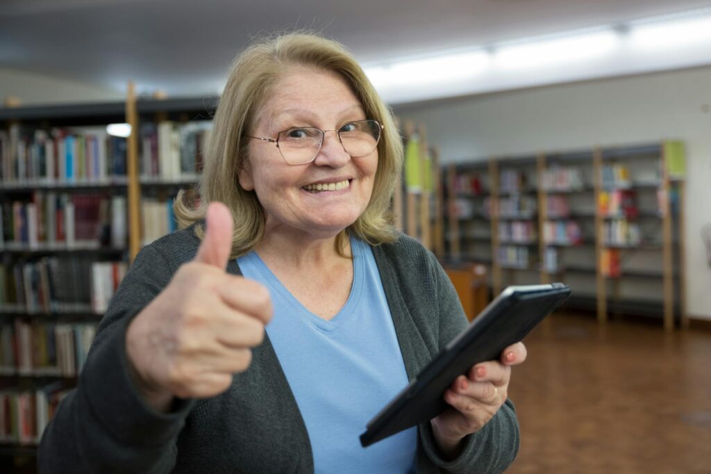 Happy senior woman gives a thumbs up while using a tablet in a library, embracing technology.
