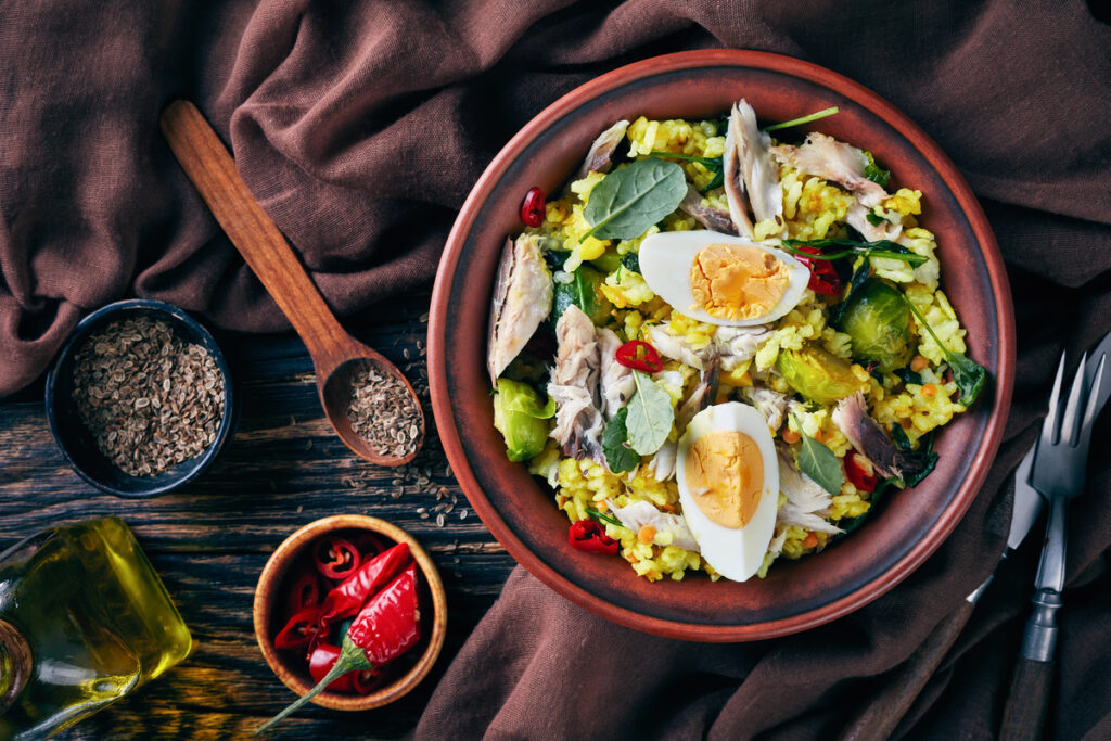 Delicious Kedgeree with flaked smoked fish, hard boiled eggs, rice, kale, brussel sprouts, spices and herbs in a bowl on an old wooden table with fennel seeds, view from above, flatlay, close-up