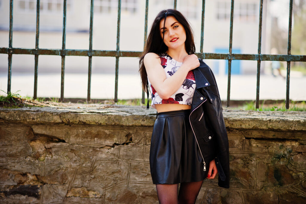 Young goth girl on black leather skirt, jacket and high heels punk shoes against iron fence.