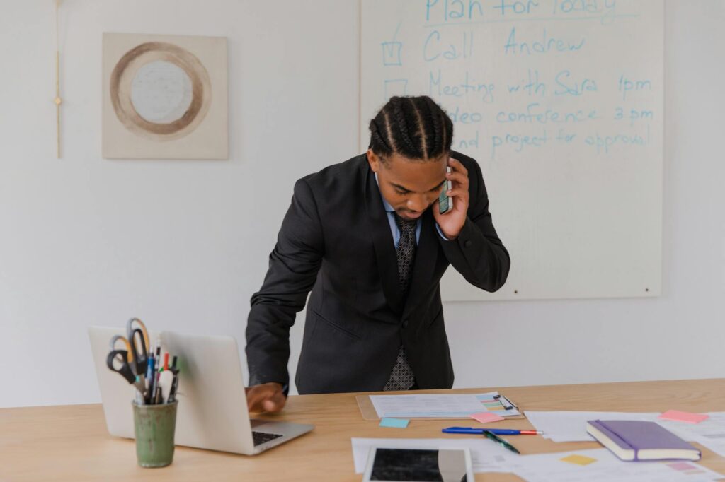 Professional businessman multitasking with phone and laptop at office desk, surrounded by documents.