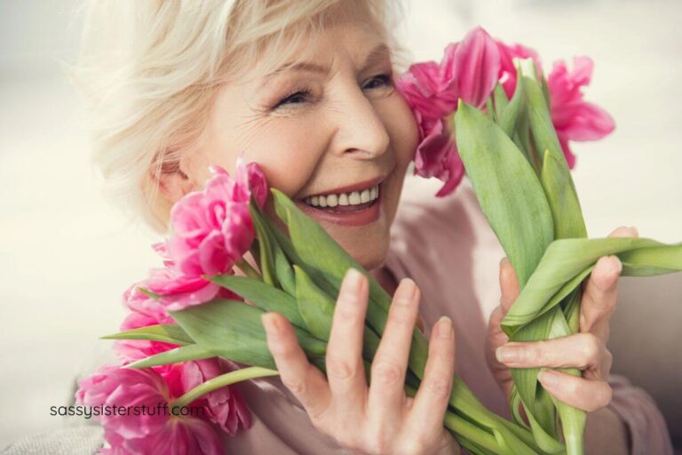senior lady holds pink flowers up to her face and smiles because she loves herself.