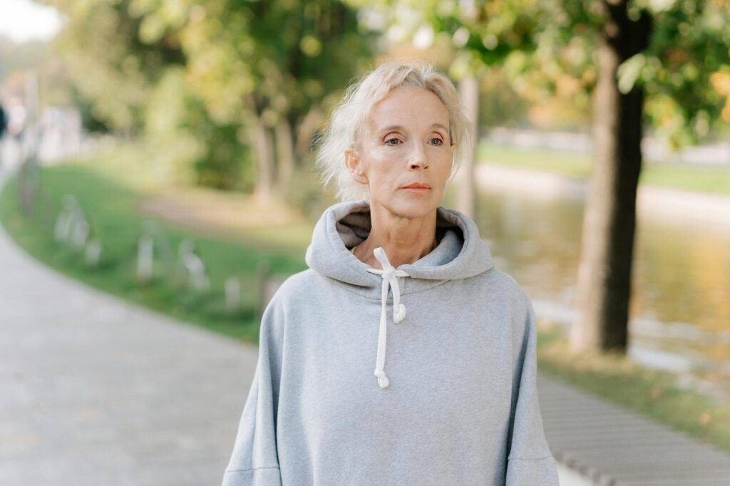 Senior woman in a hoodie strolling through a sunny park, reflecting tranquility.